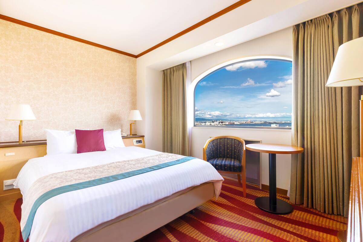 A hotel room with a large bed, two bedside lamps, a small table, a chair, and a view of the city through a large window.