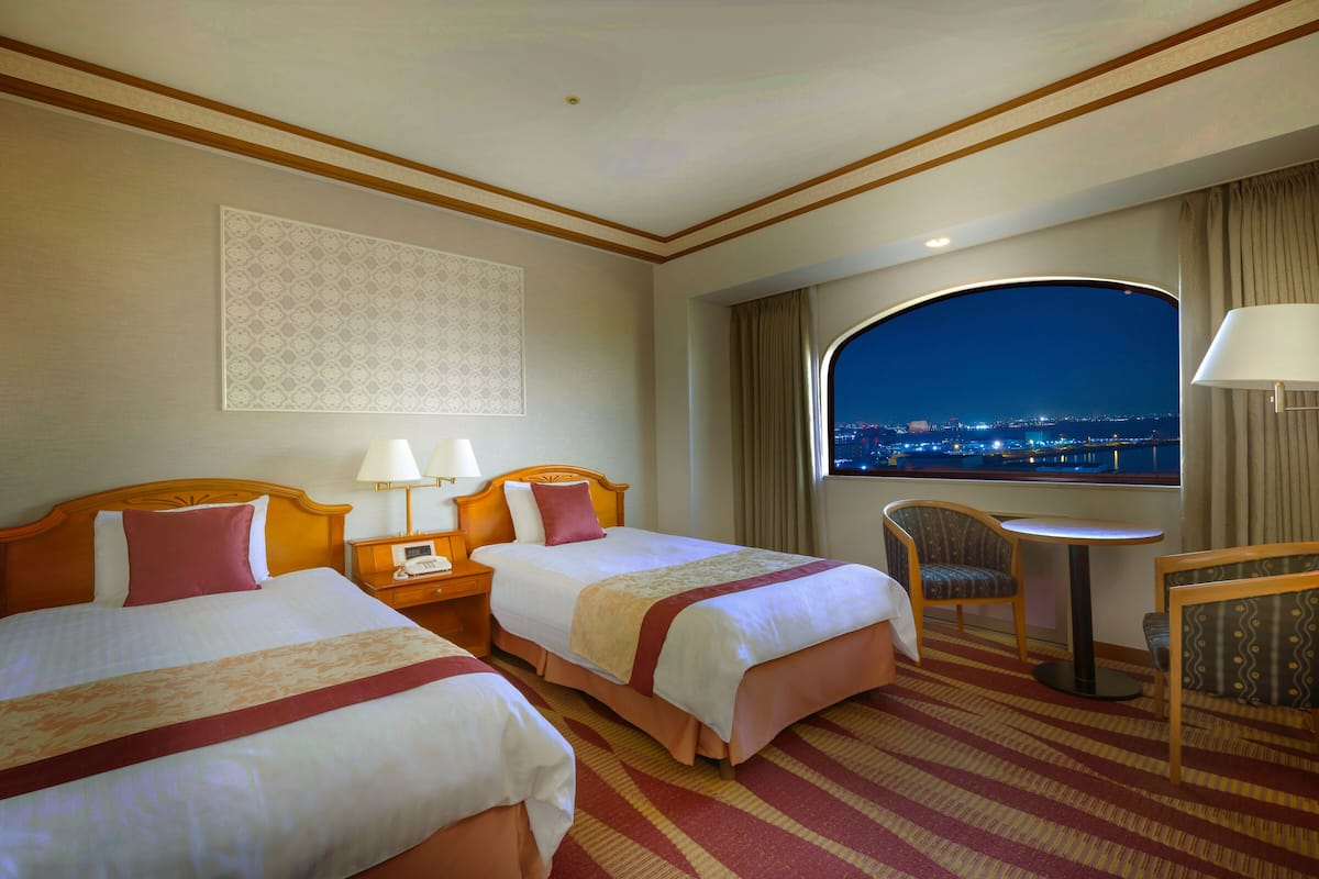 A hotel room with two beds, a nightstand, a table with a chair, and a view of the city at night.
