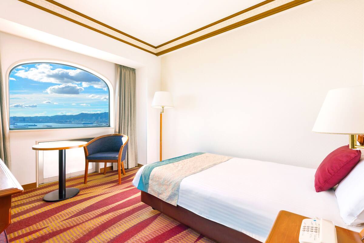 A hotel room with a large window, a bed, a desk, a chair, and a lamp.