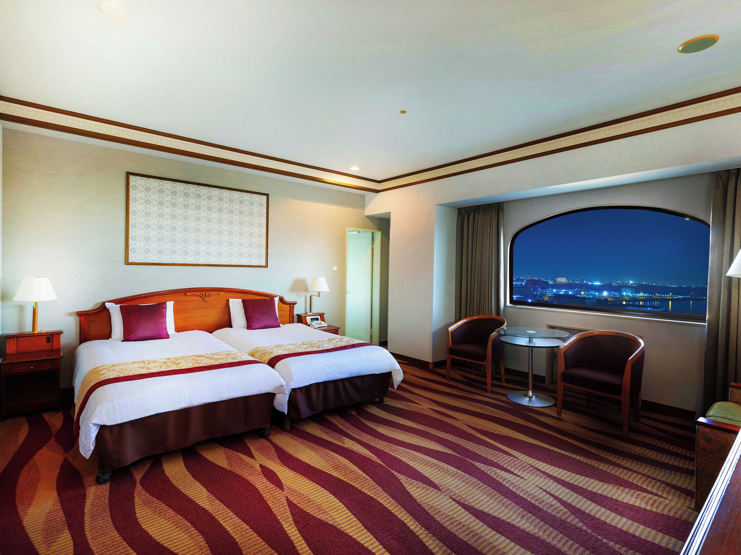 A hotel room with two beds, a desk, chairs, a view of the city at night, and a patterned carpet.