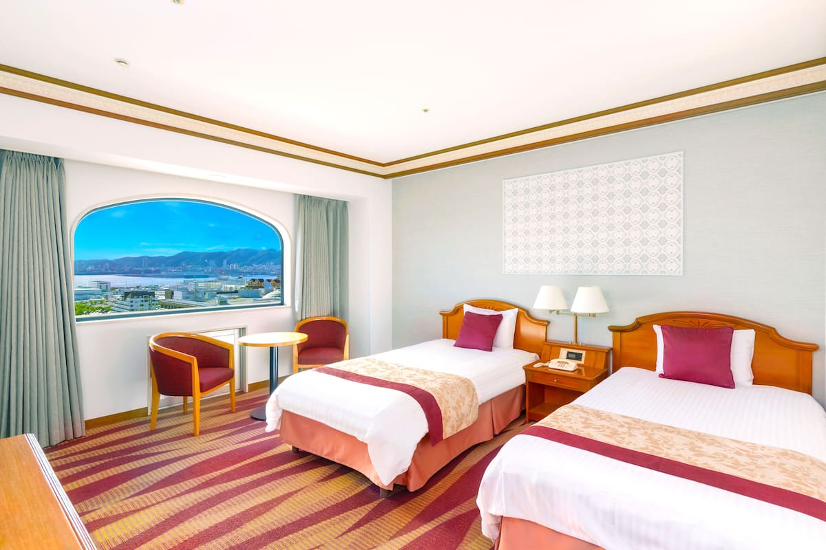 A hotel room with two beds, a view of a cityscape, a small table, and a chair.