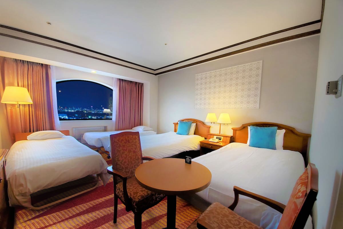 A hotel room with two beds, a small table, and a chair. There is a window with curtains, a wall-mounted lamp, and a patterned wall hanging.