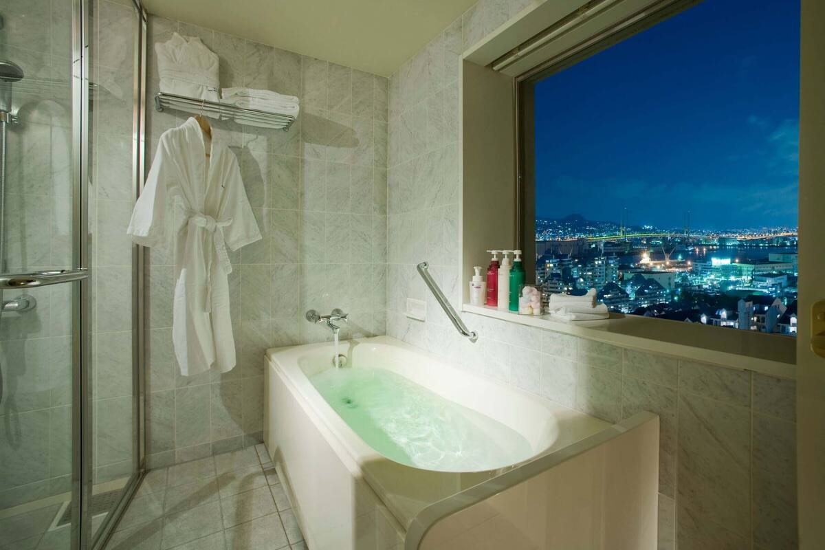A modern bathroom with a glass-enclosed shower, a freestanding bathtub, and a window offering a city view at night.