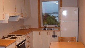 A compact kitchen with white cabinets, a stove, oven, and a refrigerator. There's a window with a view of greenery and a body of water. - Roligheden Ferieleiligheter (Kristiansand)