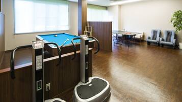 A pool table, exercise equipment, a ping pong table, and seating in a room with wooden flooring and a window with blinds.