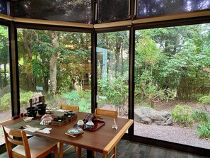 Una zona de menjador amb una taula preparada per a dues persones, vista d'un bosc a través de grans finestres i un element de pedra a l'exterior.