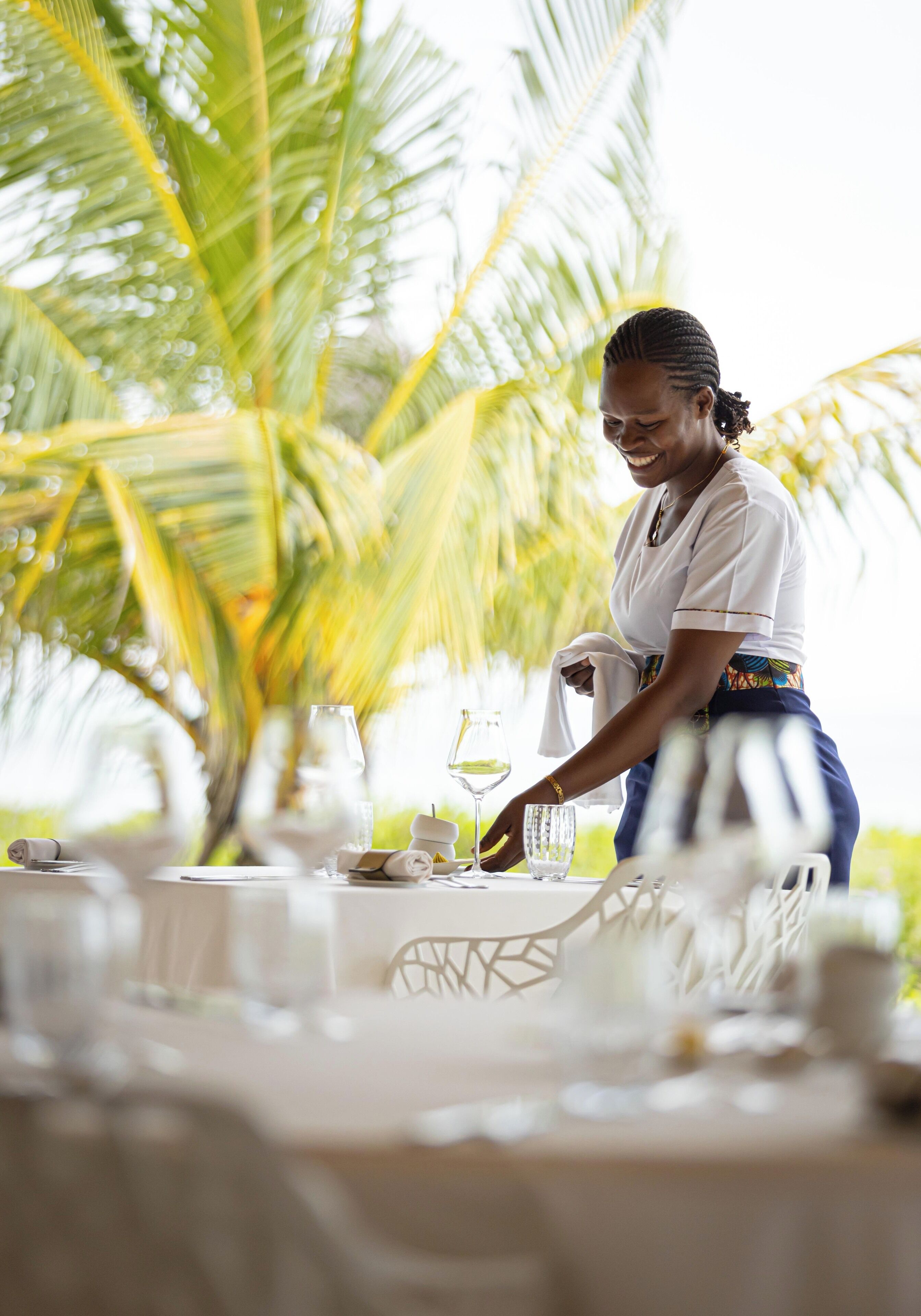 Une personne prépare une table avec des nappes blanches et de la verrerie, des palmiers en arrière-plan, dans un décor tropical.