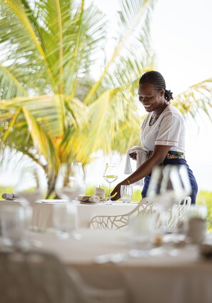 A person setting up a table with white tablecloths and glassware, palm trees in the background, and a tropical setting.