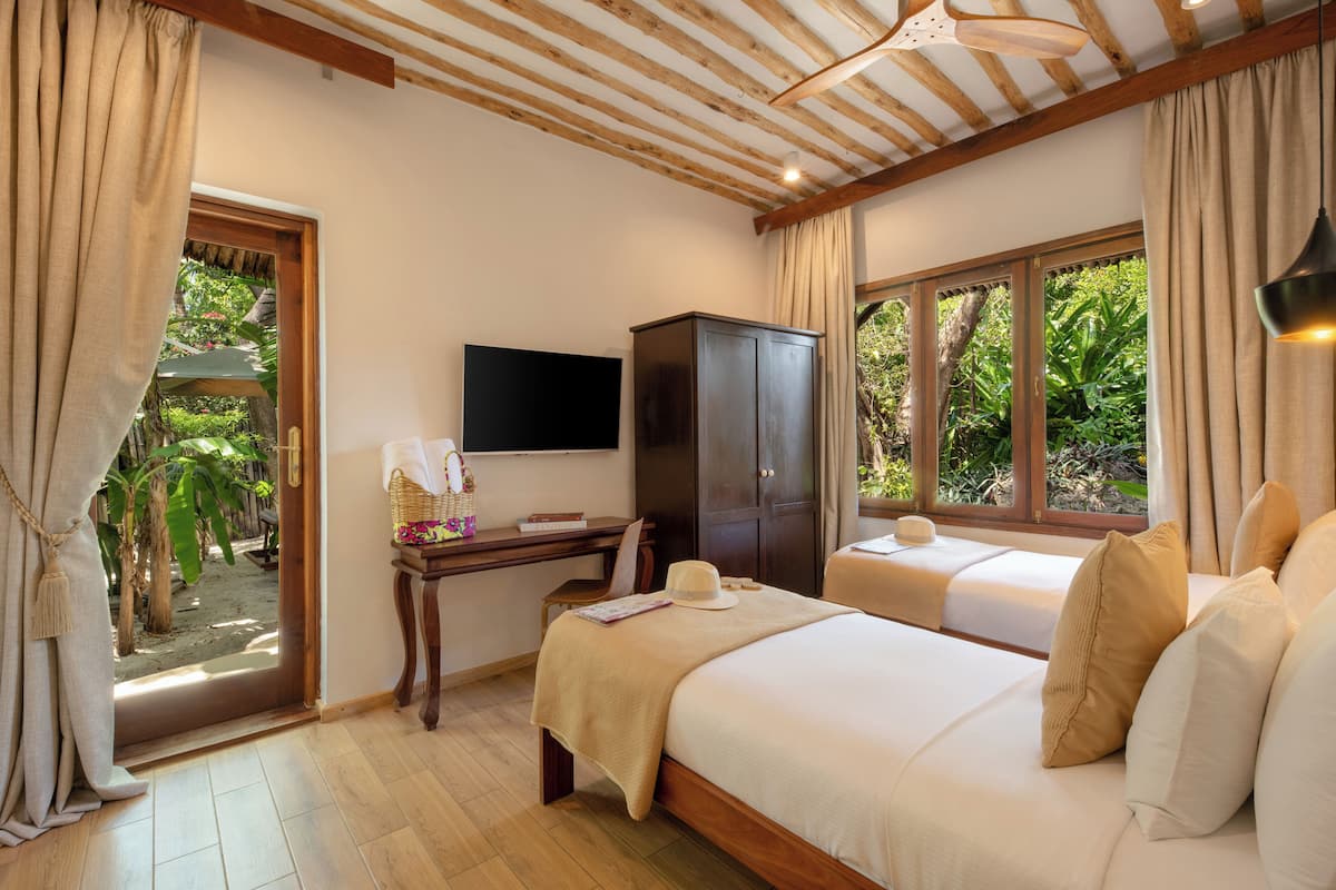 A hotel room with two beds, a desk, a TV, and a view of greenery.