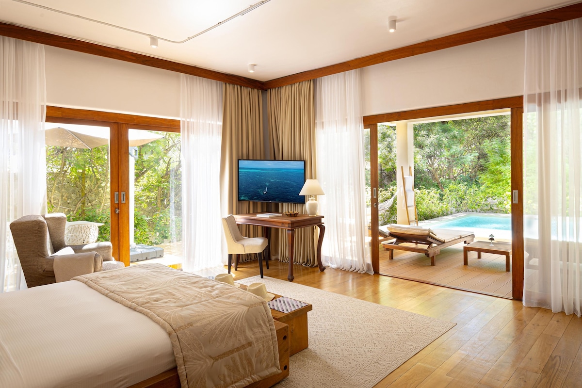 A spacious bedroom with a large bed, a desk with a TV, and a view of an outdoor pool.