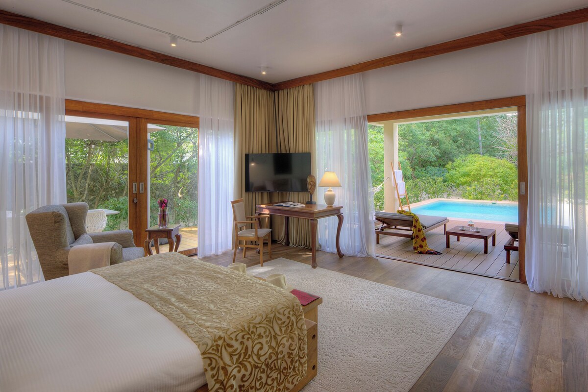 Une chambre spacieuse avec un grand lit, un bureau et une télévision. La fenêtre offre une vue sur une piscine et sur la verdure environnante.