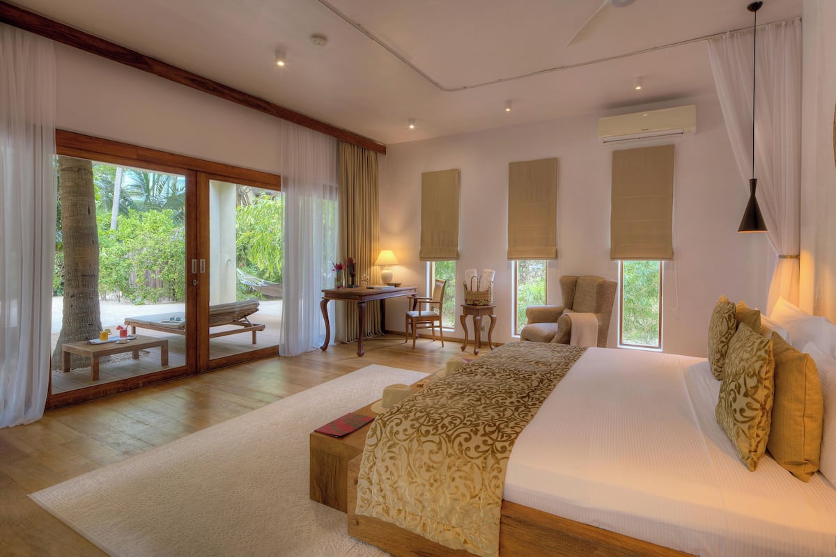 A spacious bedroom with a large bed, a desk, and a chair. There are large windows with curtains, a hammock outside, and a view of greenery.