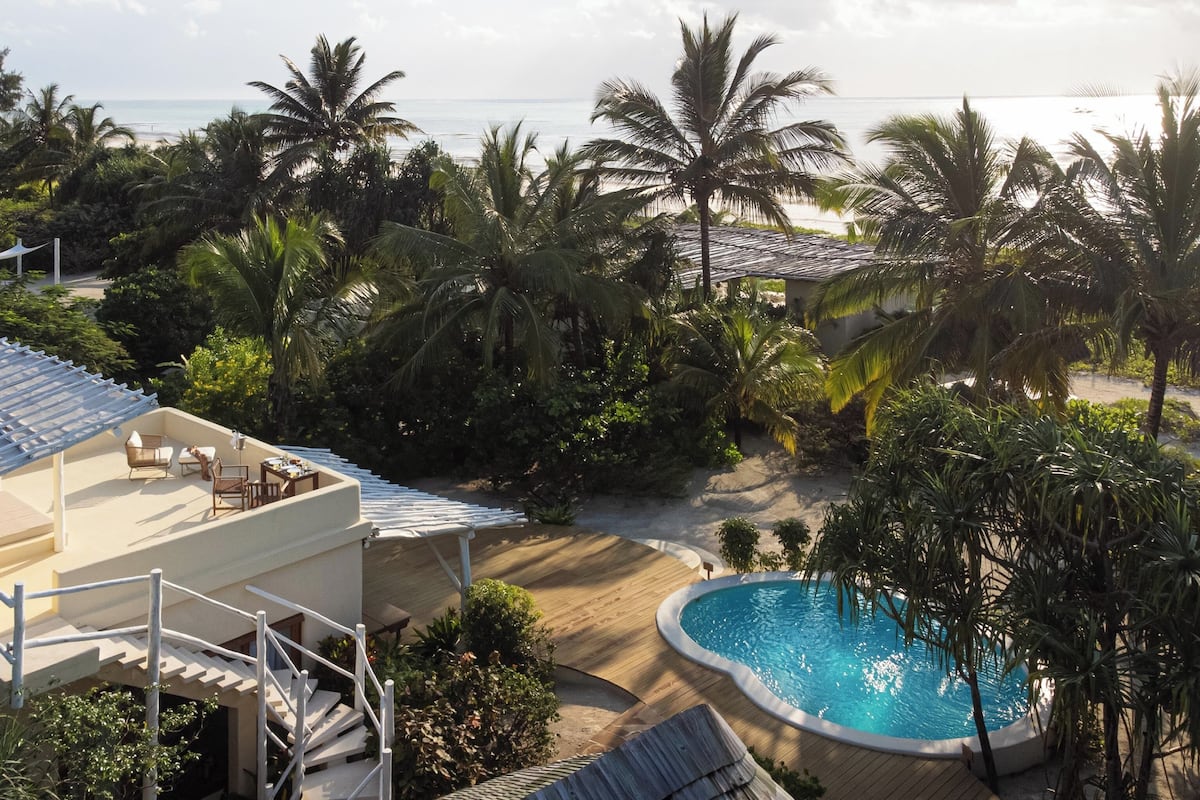 A pool area with a view of the beach, surrounded by palm trees and a building with outdoor seating.