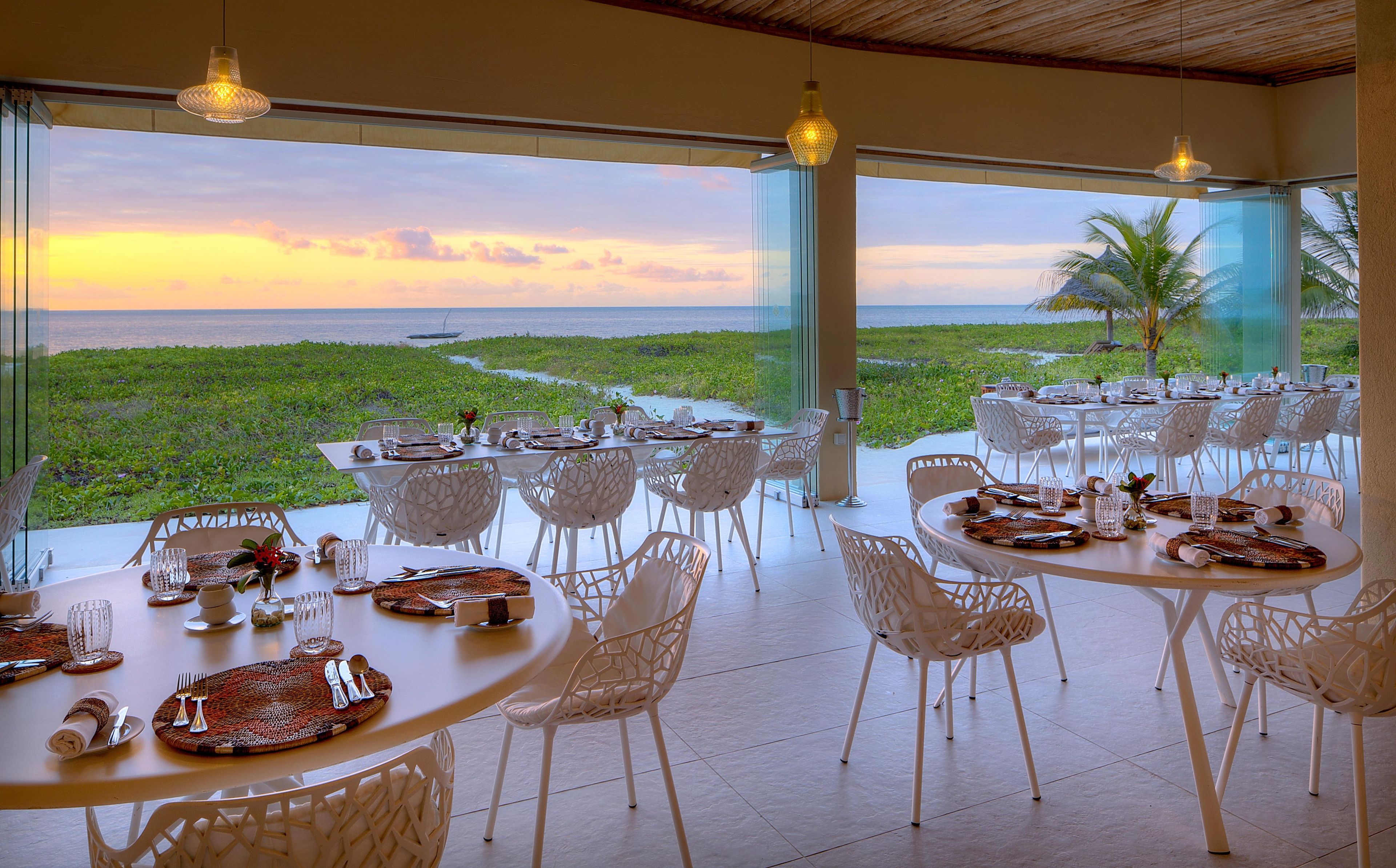 A dining area with tables set for two, overlooking a lush landscape and the sea at sunset.