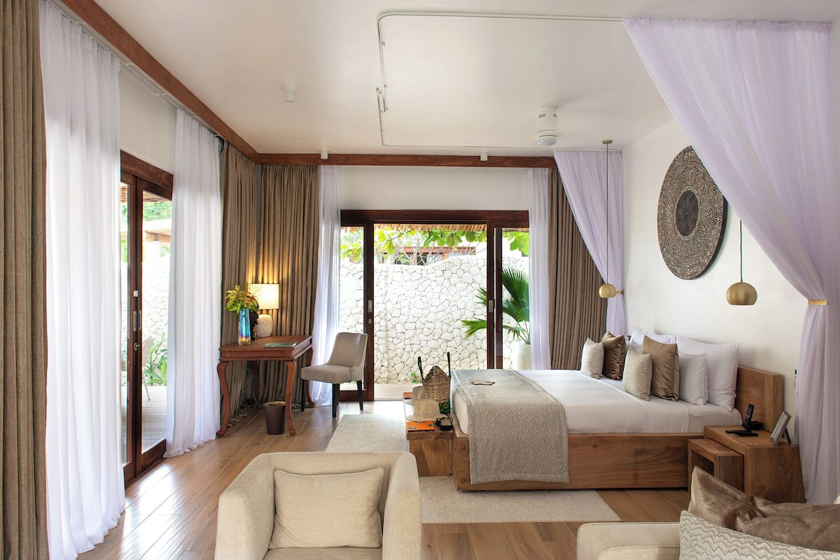 A bedroom with a large bed, a desk, and a chair. There is a stone wall and a view of greenery outside.