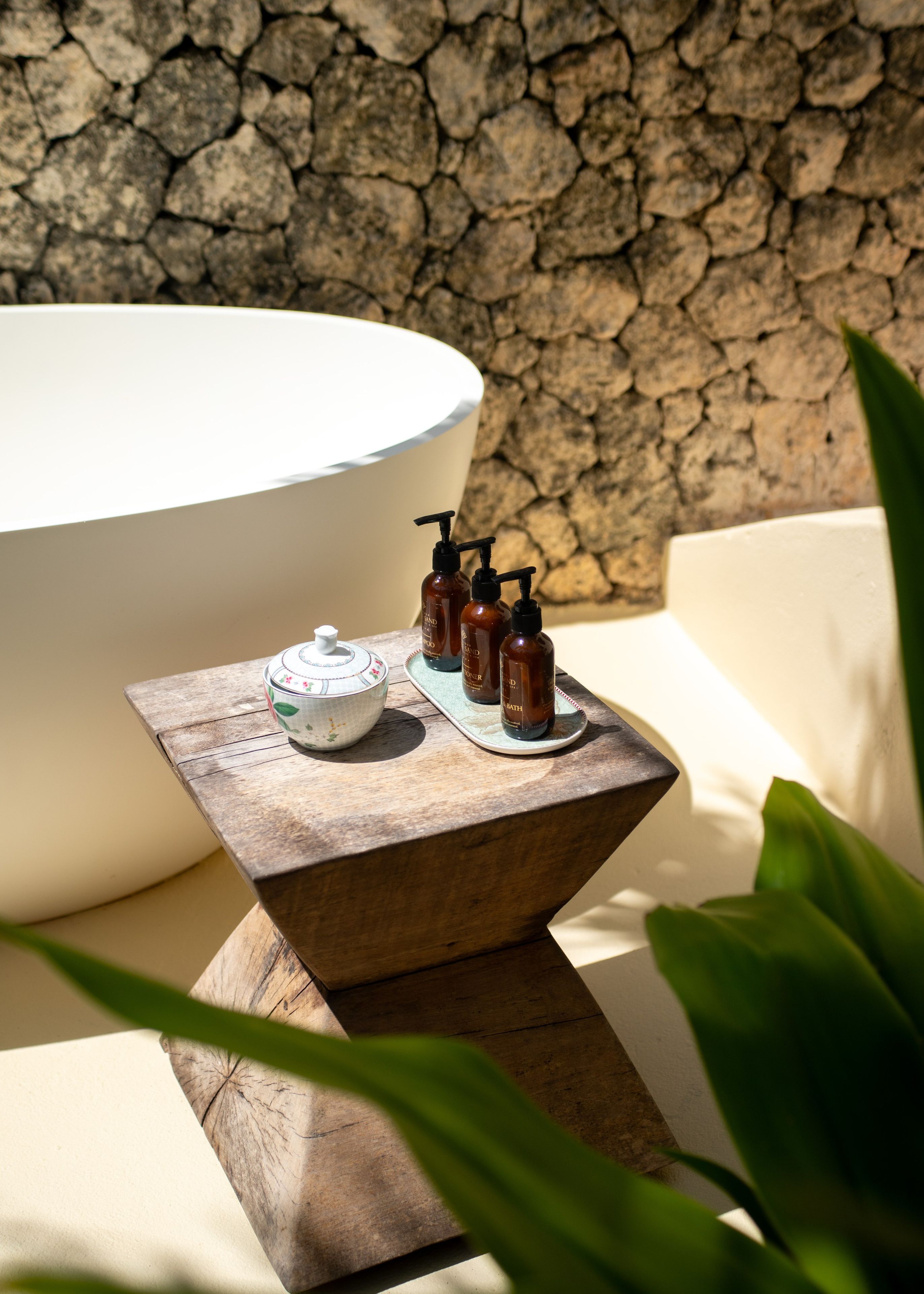 A wooden table with a teapot and bottles, a white bathtub, and a stone wall in the background.