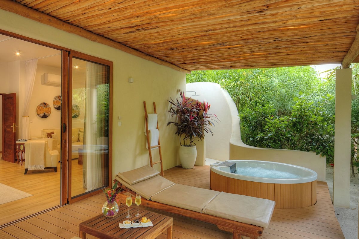 A modern hotel room with a wooden ceiling, a glass door leading to a balcony, a lounge area with a sofa and a table, and a hot tub.