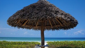 Un parasol de plage avec un toit de chaume, deux chaises longues et une plage de sable offrant une vue sur l’océan.