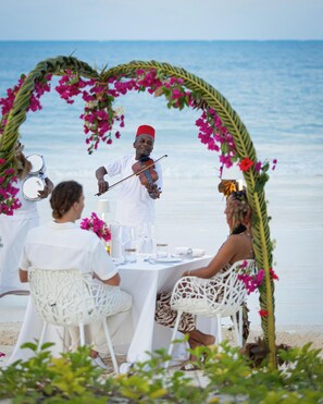 Een restaurant aan het strand met een muzikant die optreedt, omringd door gasten die aan een tafel zitten met uitzicht op zee.
