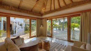 Une pièce spacieuse avec un plafond en bois, de grandes portes-fenêtres coulissantes donnant sur un espace extérieur doté d’une terrasse en bois et de plantes tropicales.