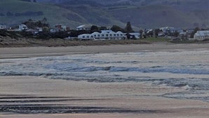 Ein Küstenblick mit einem Anwesen direkt am Strand, einschließlich eines großen Hauses und kleinerer Gebäude, mit Hügeln im Hintergrund.