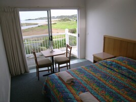 A hotel room with a bed, a small table with chairs, a balcony with a view of the beach and water, and a wooden headboard.