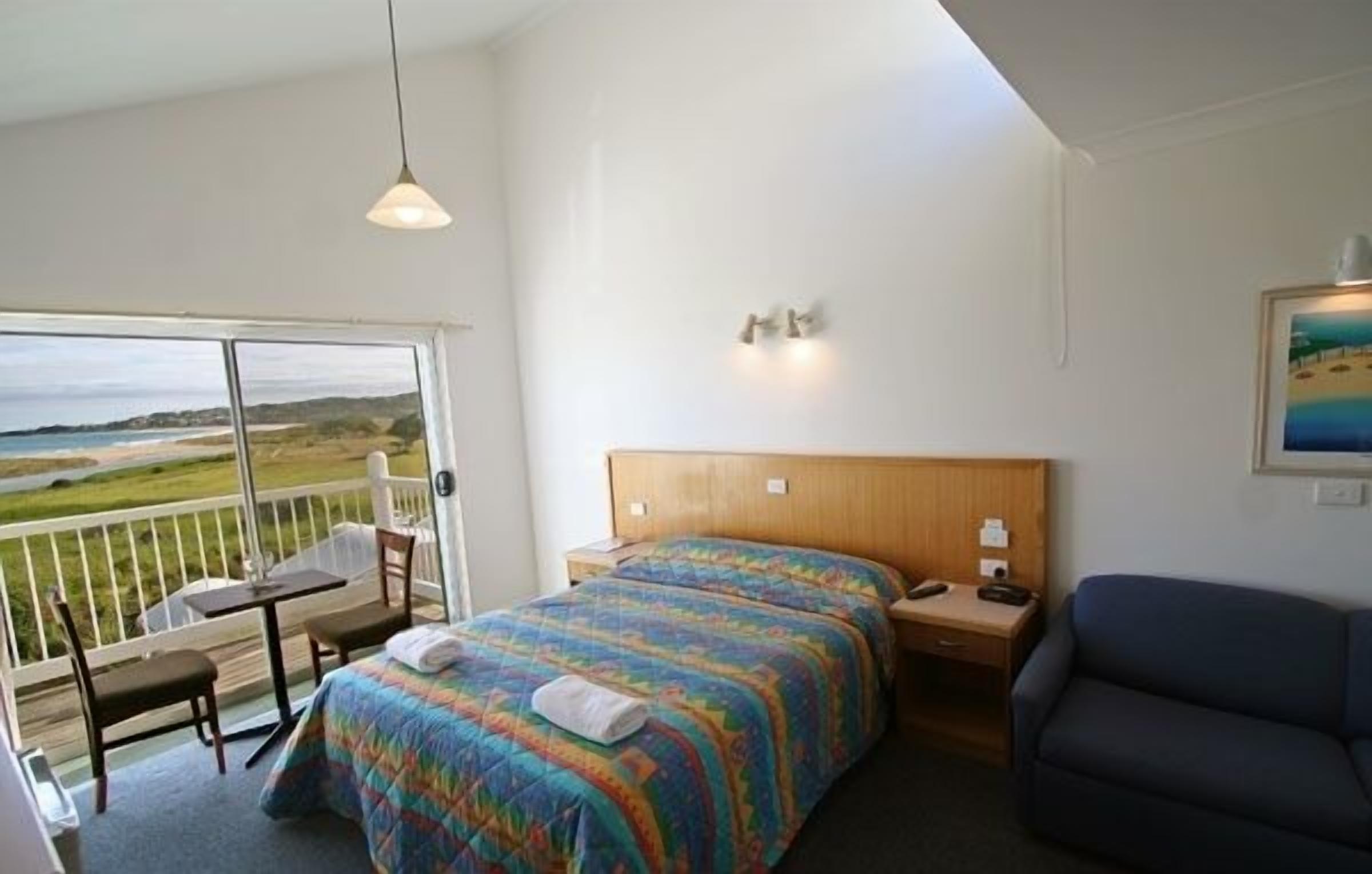 A room with a bed, a desk, a chair, a sofa, and a balcony with a view of the beach.
