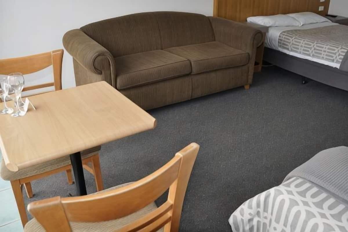 A room with a wooden table, two wooden chairs, a brown sofa, and a bed with a grey patterned bedspread.