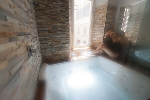 A modern bathroom with a stone wall, a large bathtub, and a window providing a view of buildings outside. - Guesthouse o Affittacamere Magenta Pantheon Navona (Rome)