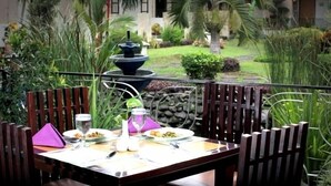 Una mesa de comedor al aire libre con sillas, una fuente de agua y abundante vegetación de fondo.