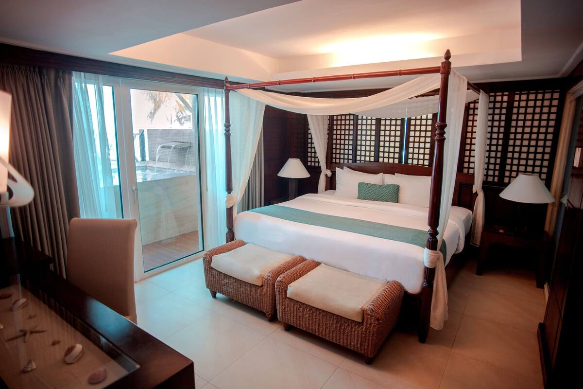 Un dormitorio amplio con cama con dosel, una zona de estar con mesa y vistas a una piscina exterior.