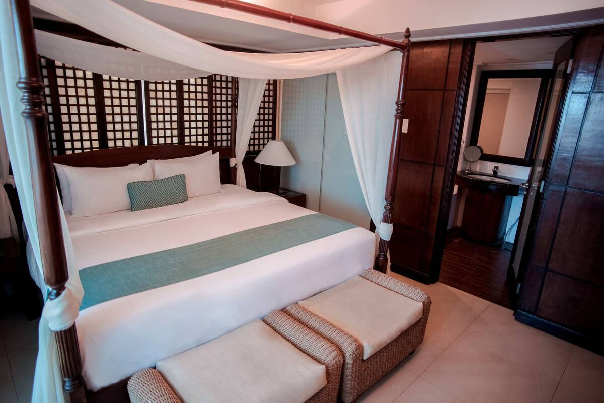 Una cama con dosel y cuatro columnas, con una manta verde, una mesita de noche con lámpara y un armario de madera con espejo.