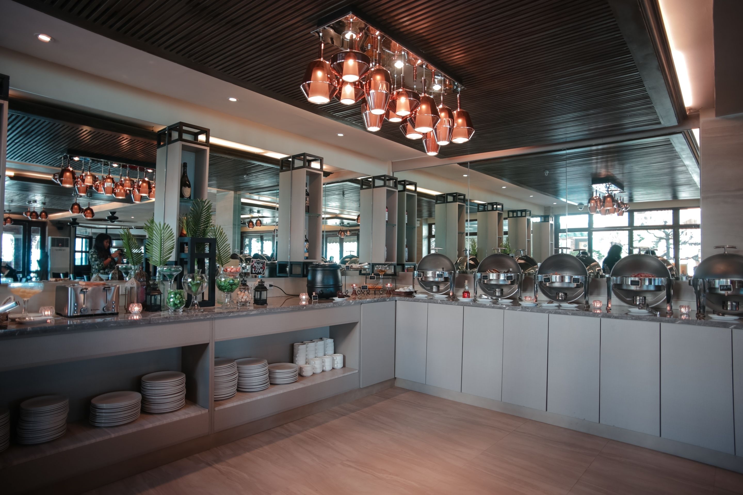 A modern kitchen with copper pendant lights, a large island with various kitchenware, and a view of the city through large windows.
