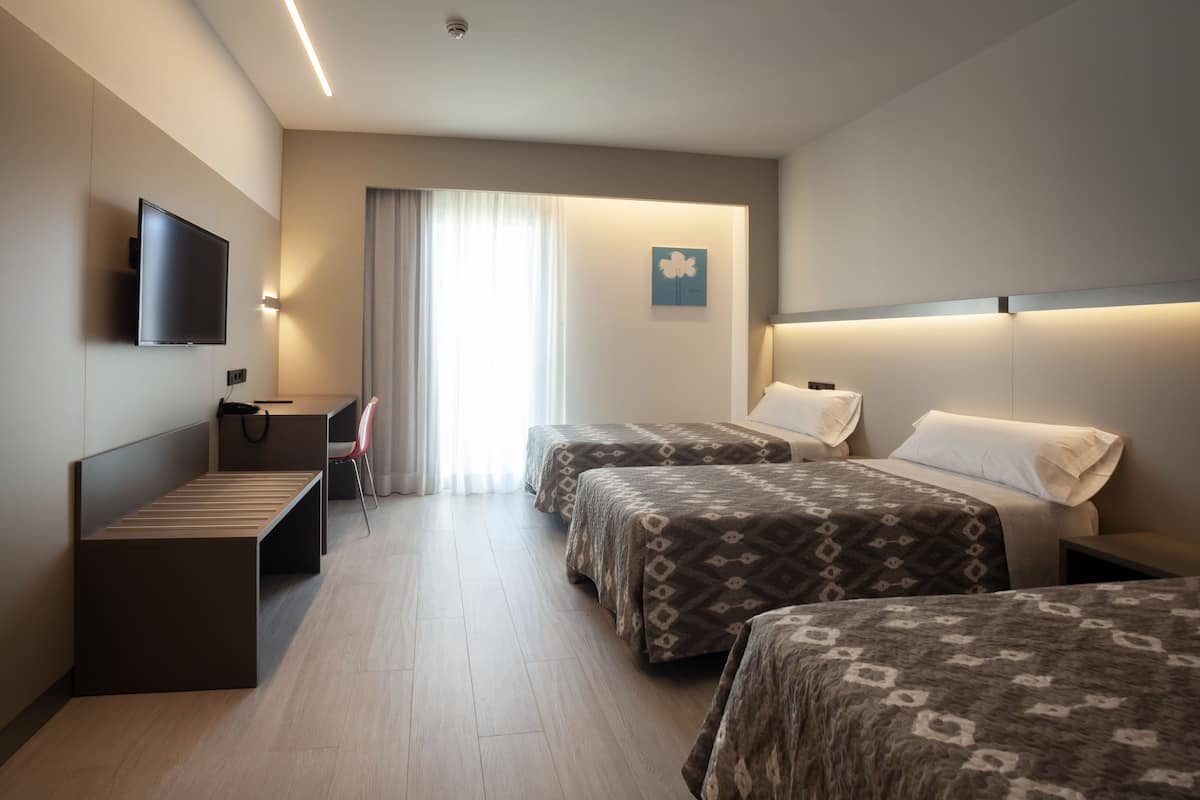Quarto de hotel com duas camas, uma escrivaninha, uma cadeira, televisão e janela com cortinas.
