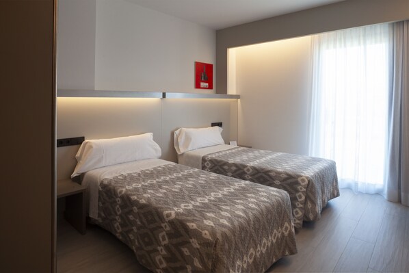 Habitación de hotel con dos camas, una mesita de noche, una lámpara de pared y un ventanal con cortinas.
