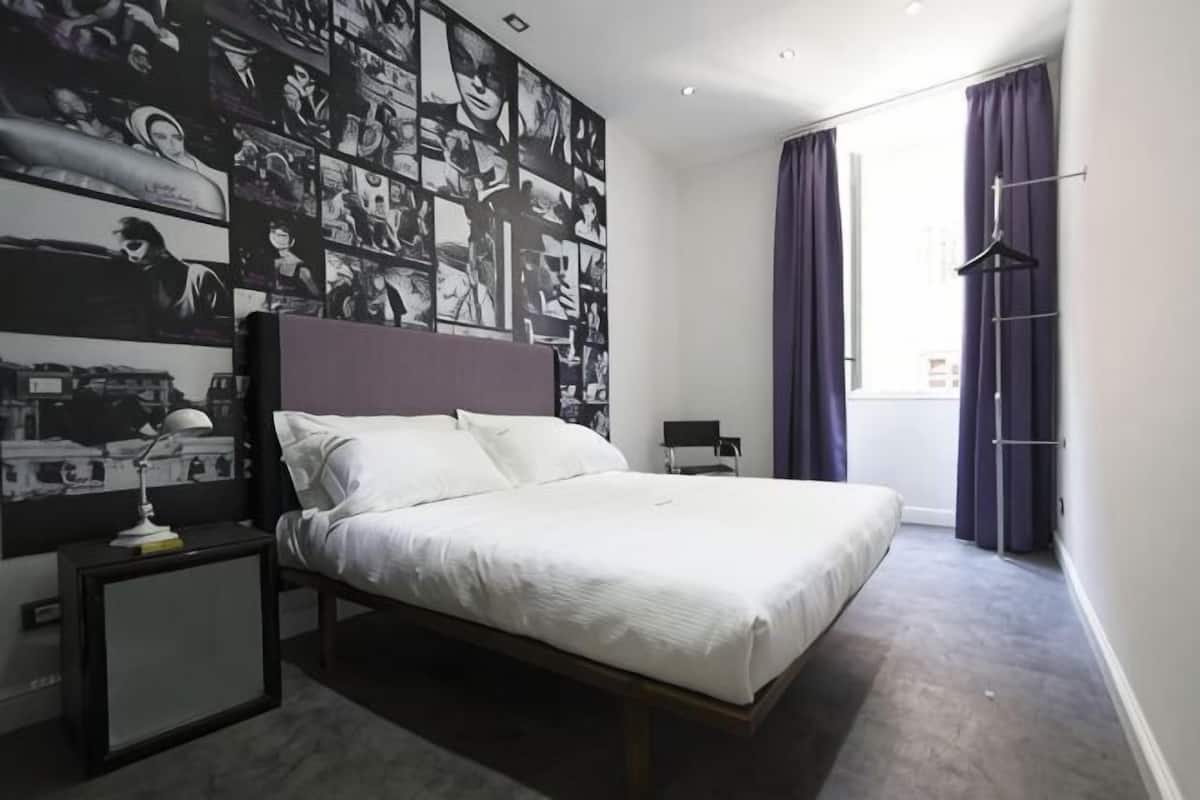 A bedroom with a bed, a nightstand, a wall with photos, a window with curtains, and a hanging coat rack.