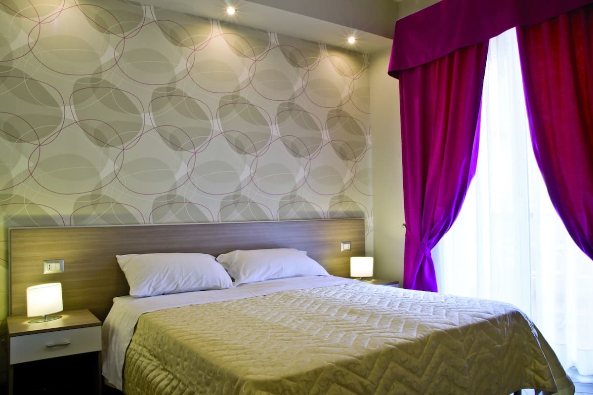 Una habitación de hotel con cama, mesitas de noche y ventana con cortinas.