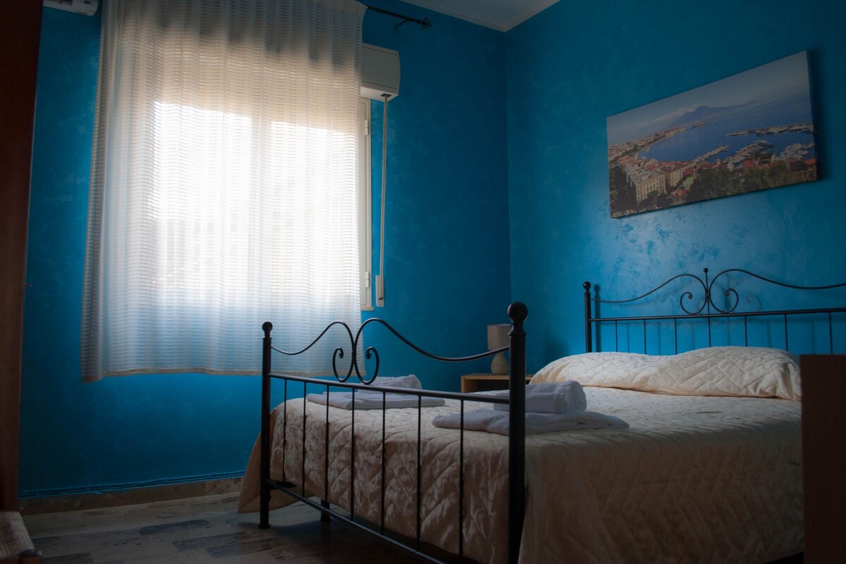 A bedroom with a bed, a window with sheer curtains, and a painting on the wall.