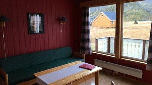 A room with a green sofa, a wooden table with a laptop, a window with a view of a house and mountains, and a radiator. - Nordkapp Camping (Nordkapp)