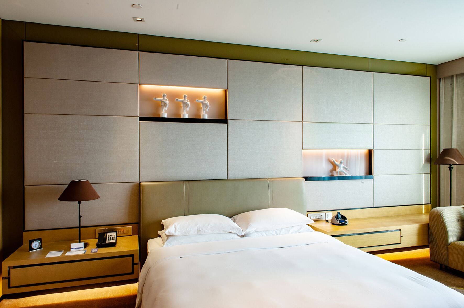 A hotel room with a large bed, two bedside tables, a nightstand, a desk, and a chair. There are wall-mounted shelves with decorative items.