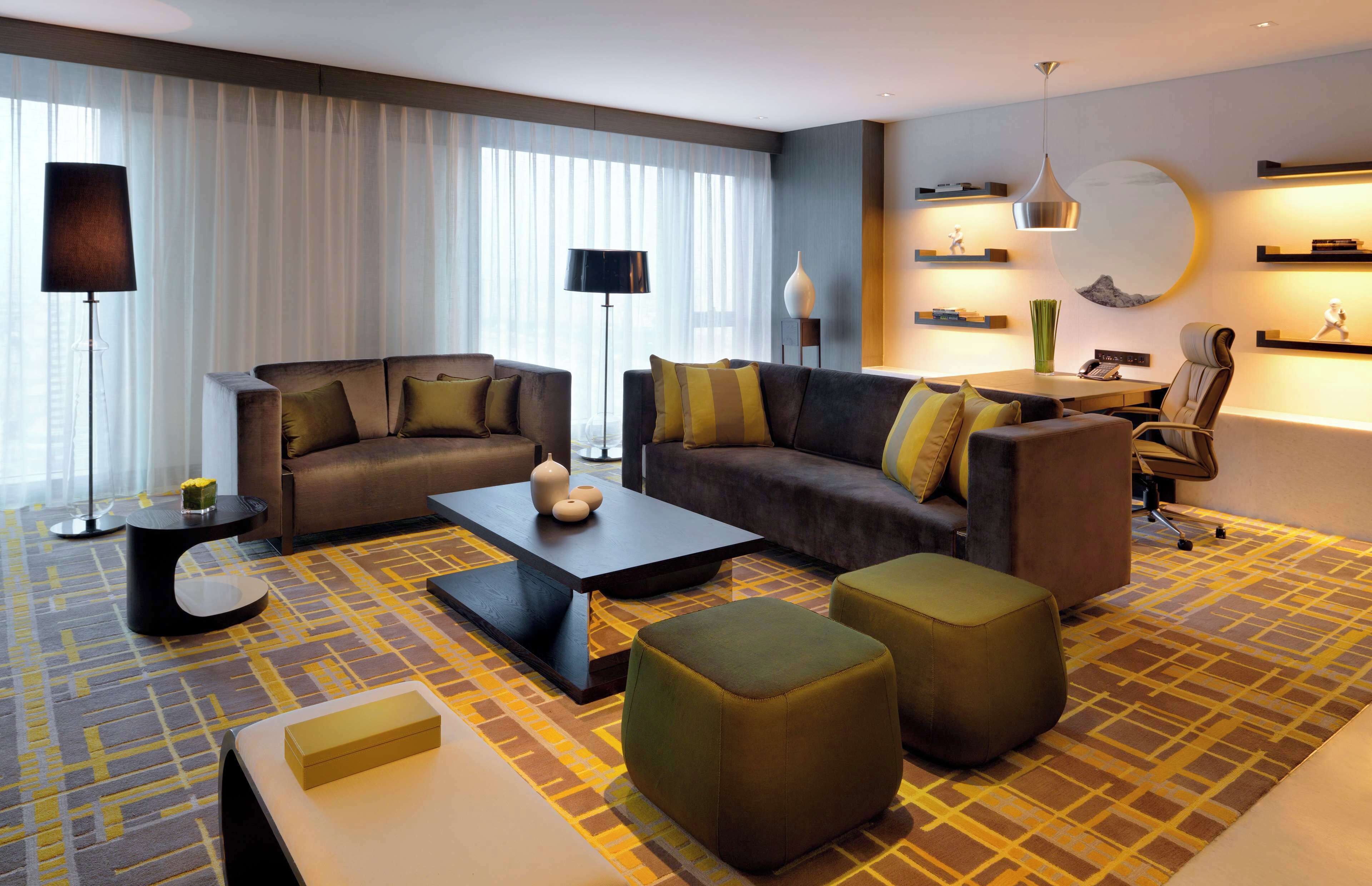 Ein modernes Hotelzimmer mit einer Sofagarnitur, einem Couchtisch, Hocker und einem Schreibtisch mit Stuhl.