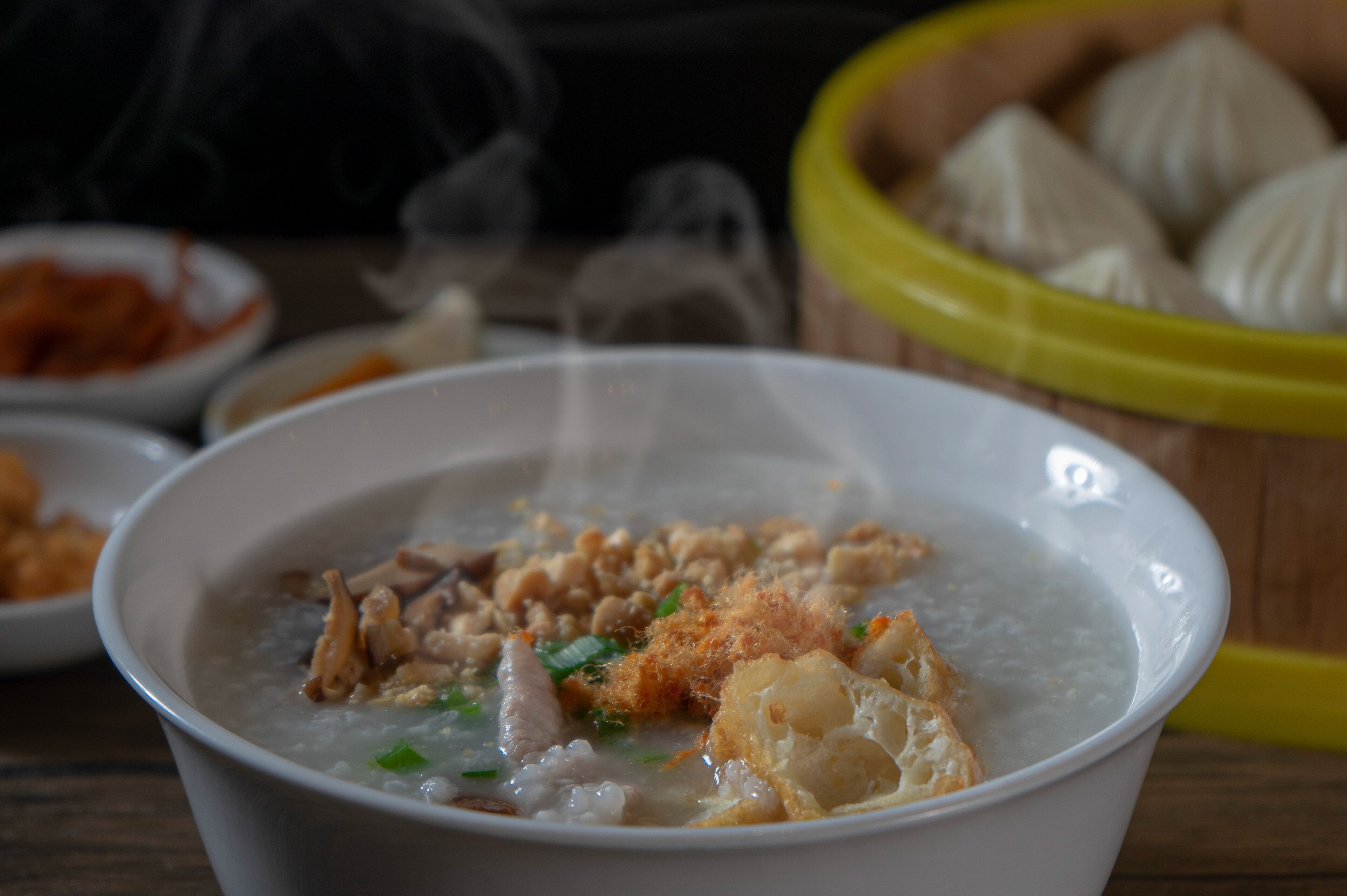 A bowl of porridge with various toppings, a basket of steamed buns, and a bowl of sauce in the background.