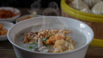 A bowl of porridge with various toppings, a basket of steamed buns, and a bowl of sauce in the background.