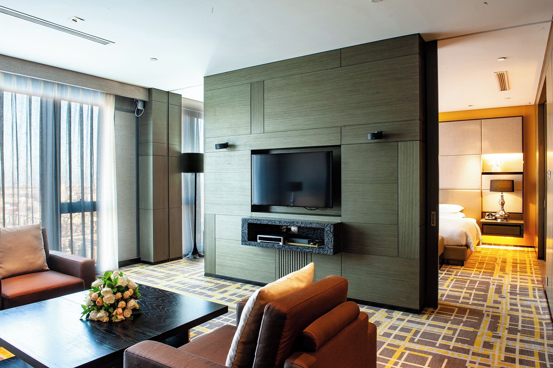 Une chambre d’hôtel moderne dotée d’un téléviseur à écran plat fixé sur un mur en panneaux de bois, d’un canapé, d’une table basse agrémentée d’un arrangement floral et d’un tapis à motifs.