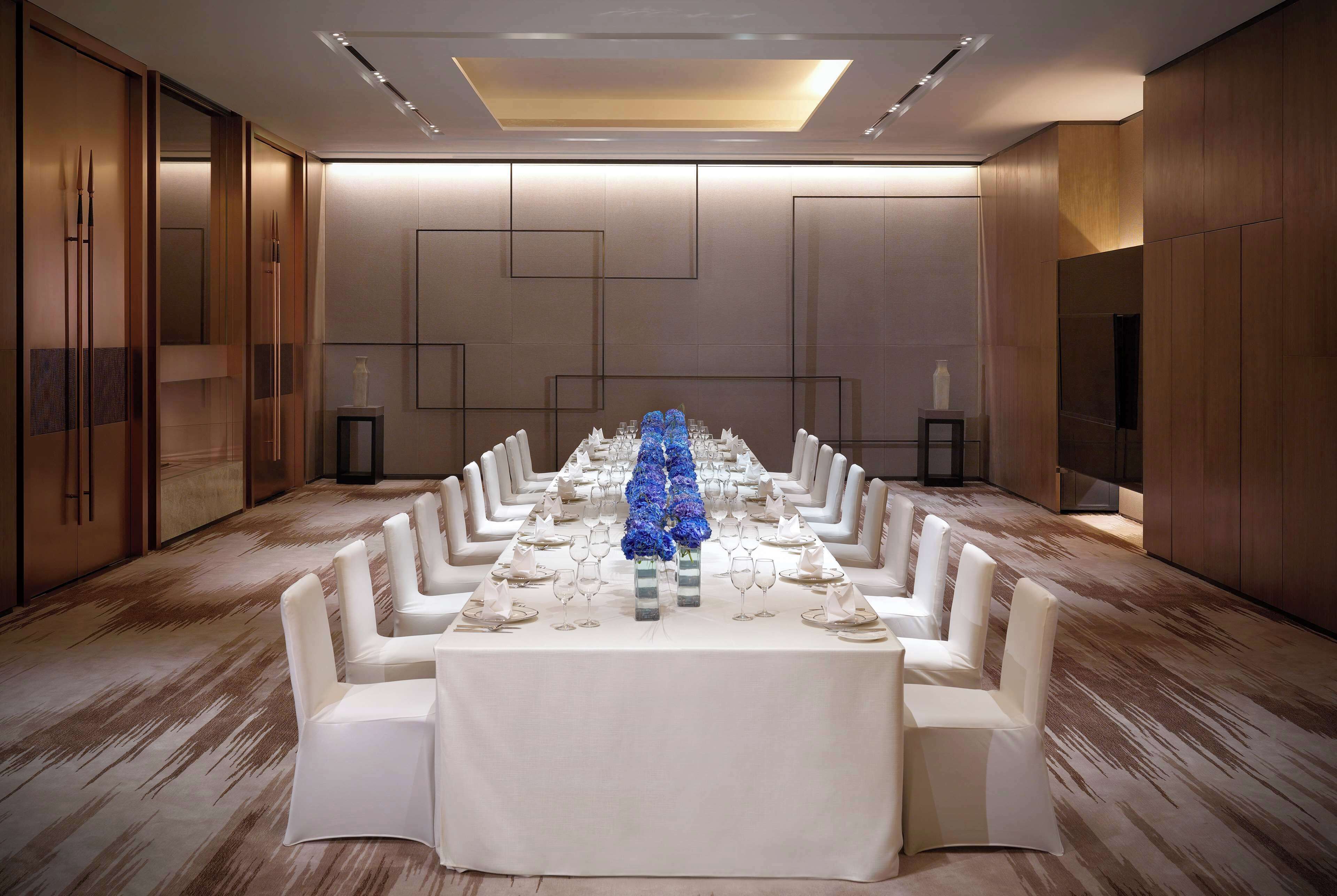 Une salle de conférence avec une longue table dressée pour un événement officiel, agrémentée de chaises blanches et d’une composition florale centrale.