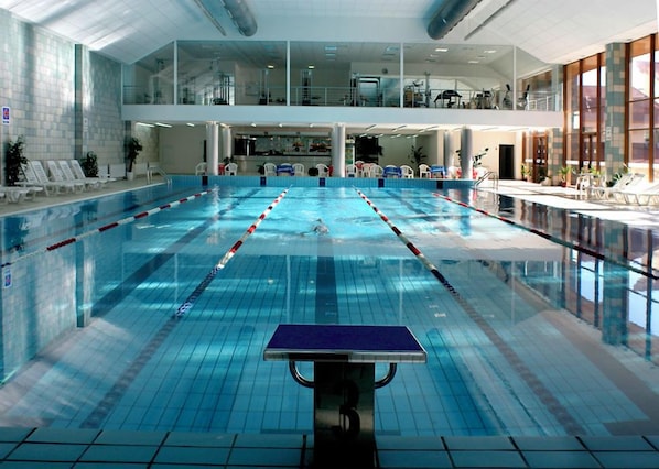 Ein großer, überdachter Schwimmbereich mit mehreren Bahnen, einer Lounge mit Liegestühlen und Blick ins Freie durch große Fenster.