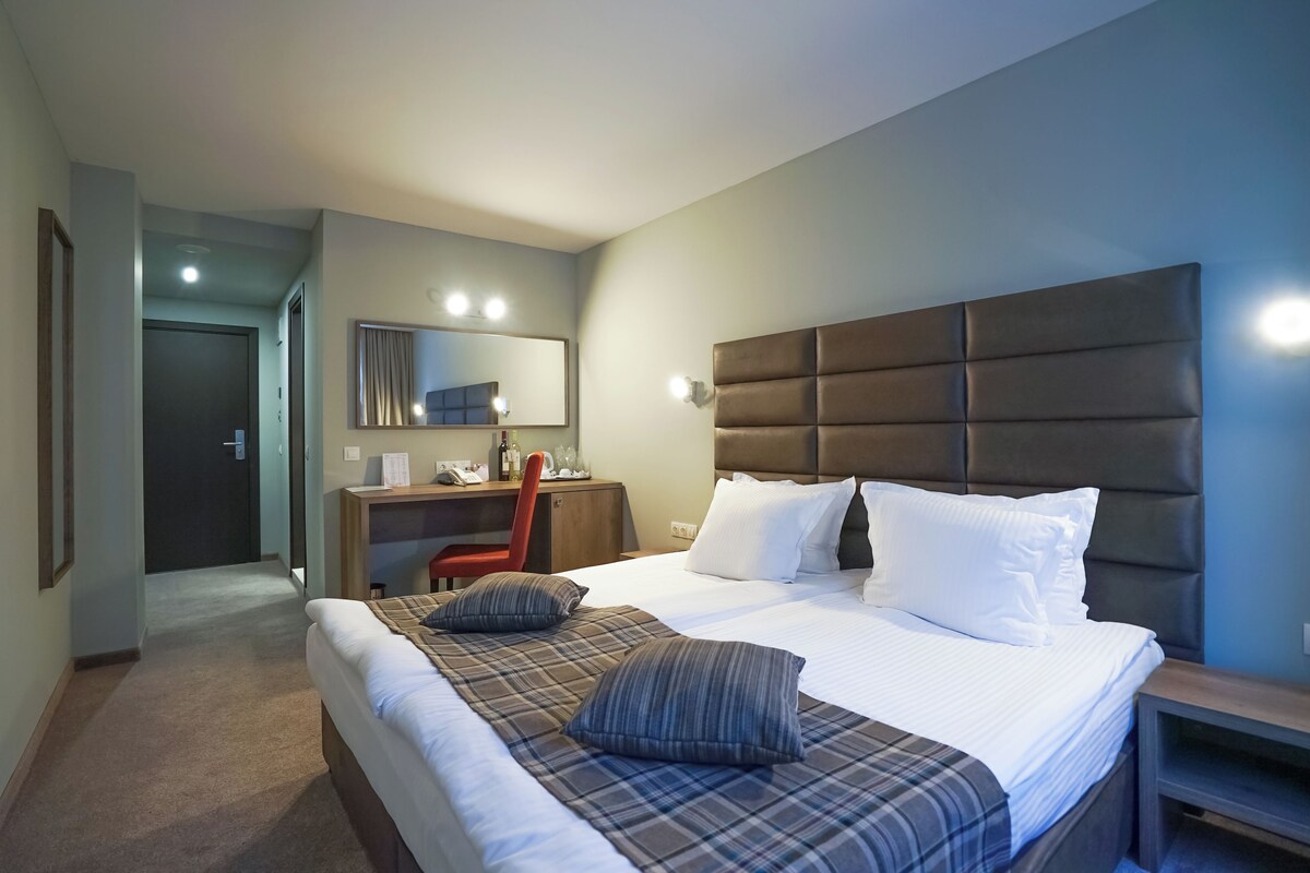 Ein Hotelzimmer mit einem großen Bett, einem Schreibtisch mit Stuhl, einem Spiegel und einer Tür zu einem anderen Raum.