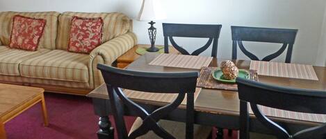 A dining area with a table set for four, surrounded by four chairs, a sofa with patterned cushions, and two framed pictures on the wall.