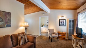 A cozy living room with a brown leather sofa, a white armchair, a wooden coffee table, and a television on a stand. A hallway leads to a bedroom with a bed and a nightstand. - Redwood Suites (Ferndale)