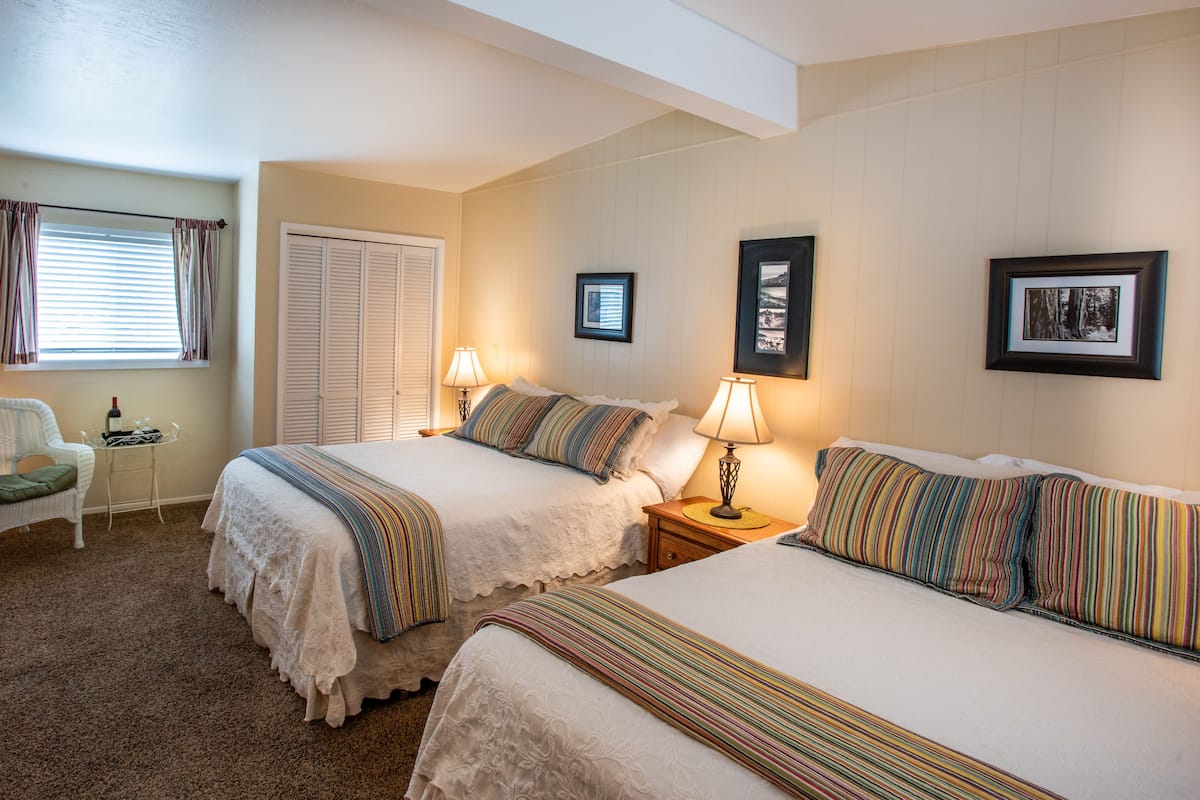 A hotel room with two beds, a chair, a small table with a bottle, and framed pictures on the wall.